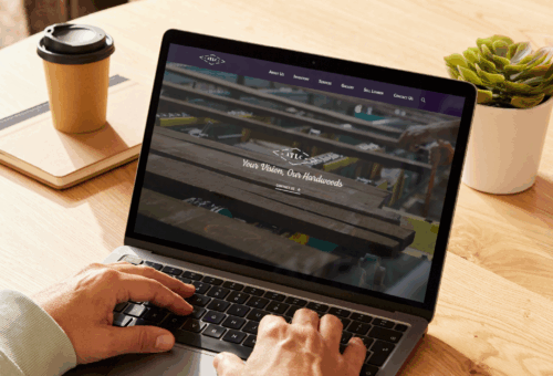 A person using a laptop displaying the Anderson Tully website homepage, highlighting the hero video section with the tagline "Your Vision, Our Hardwoods" in a real-world setting.