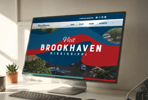 Desktop monitor on a modern workspace displaying the Brookhaven Tourism website homepage, featuring bold red and blue graphics and navigation links for Eat, Play, Stay, and Events.